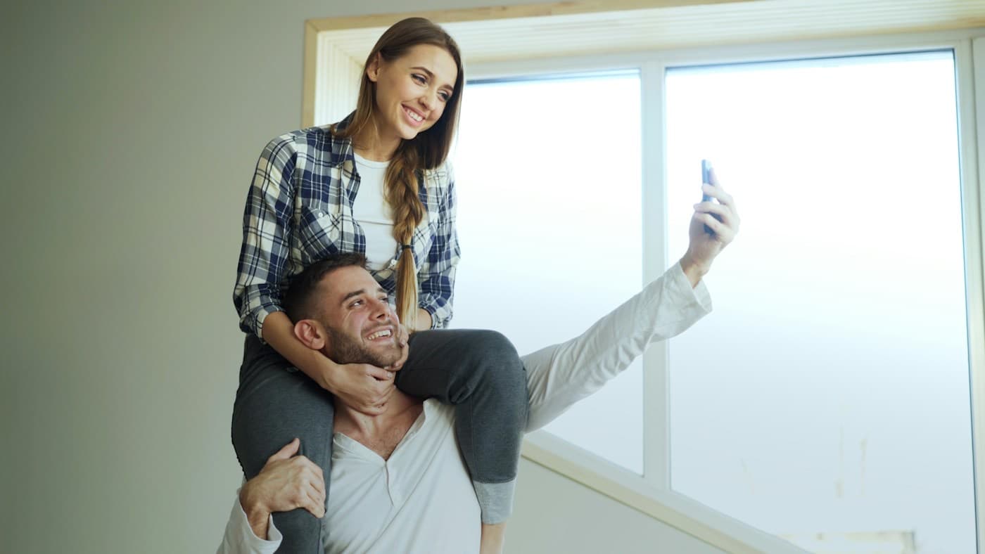 Casal sorrindo ao tirar uma selfie perto da janela de casa