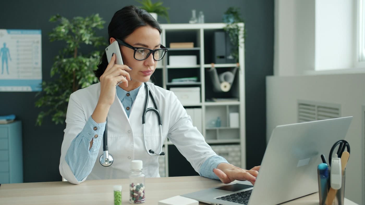 Médica de jaleco atendendo pelo telefone em frente ao notebook